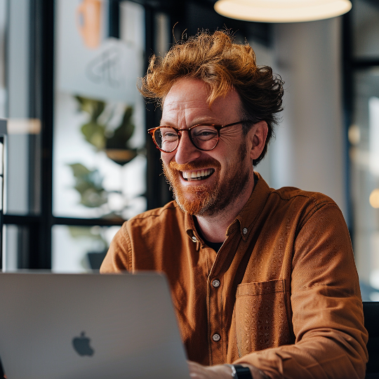 Um homem de óculos está sorrindo enquanto usa um laptop da Apple.