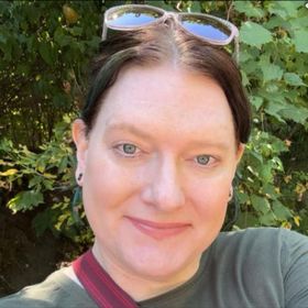 Woman with glasses atop head, smiling in front of green foliage. Wearing a green shirt and a red strap across her body.