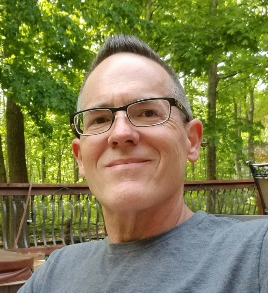 Man with glasses smiles outdoors, with trees in the background.