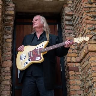 Man with long hair holding a cream-colored electric guitar, standing in a doorway with brick walls.