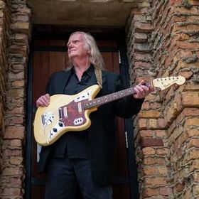 Man with long hair holding a cream-colored electric guitar, standing in a doorway with brick walls.