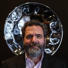 Man with beard in front of a steelpan. He wears a suit jacket and a pink patterned shirt.