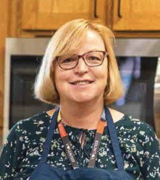 Woman with glasses and blonde hair wearing apron, smiling in kitchen setting.