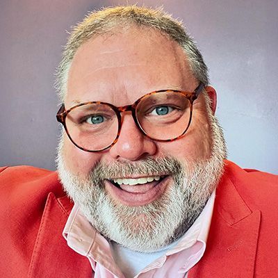 Woman with short blonde hair smiles, wearing an orange sweater and a gold necklace.
