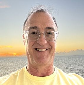 Man with glasses smiling in front of a sunset over the ocean.