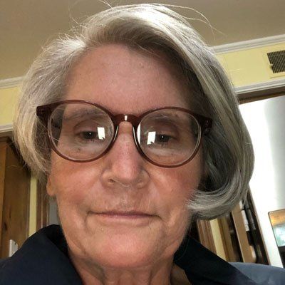 Woman with gray hair and glasses looks directly at the camera.