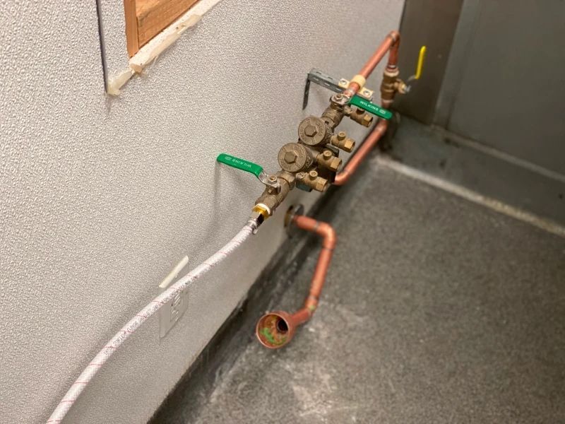 Copper pipes and valves against a gray wall, near a textured white surface, with a green-handled valve.