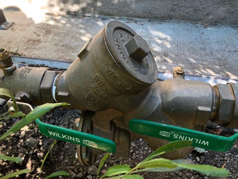 A backflow preventer with green-handled valves and a brass body sits in a garden setting.