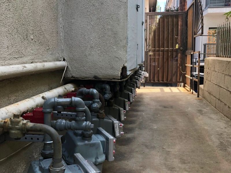 Gas meters and pipes along a concrete walkway beside a building, leading to a gate.