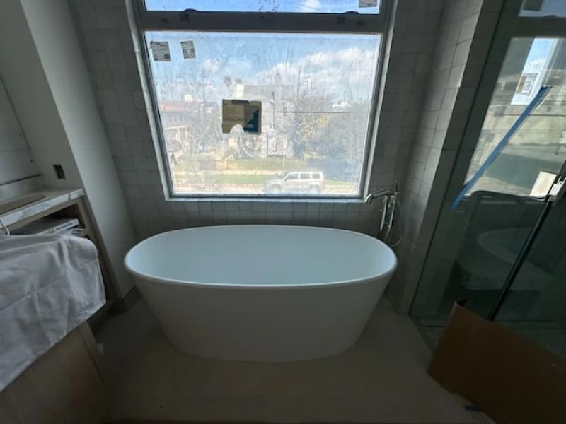 White, oval bathtub in a bathroom, beneath a large window with a view of a street.