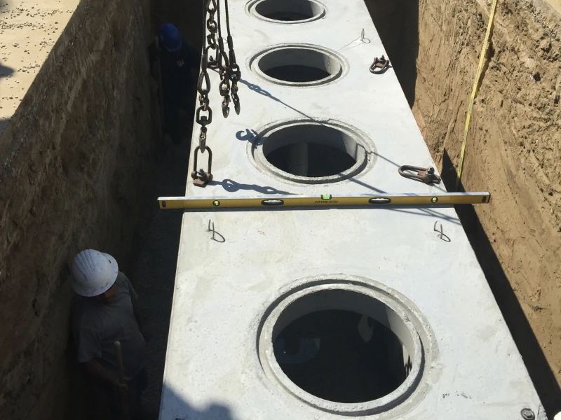 Concrete structure being lowered into a trench, workers nearby with level and chains.