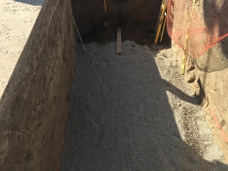 Gravel-lined trench at a construction site, with exposed earthen walls and orange safety netting.