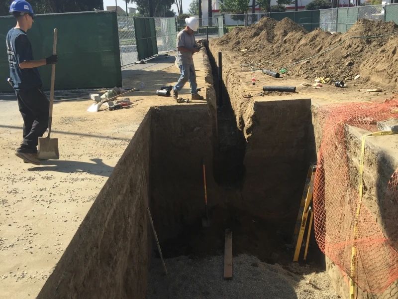 Construction workers in a trench; one with a shovel, another walking. Dirt piles, safety netting, and fencing surround.