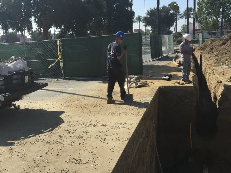 Two workers in construction area, one with a shovel, one looking at the trench. Outside, sunny day.