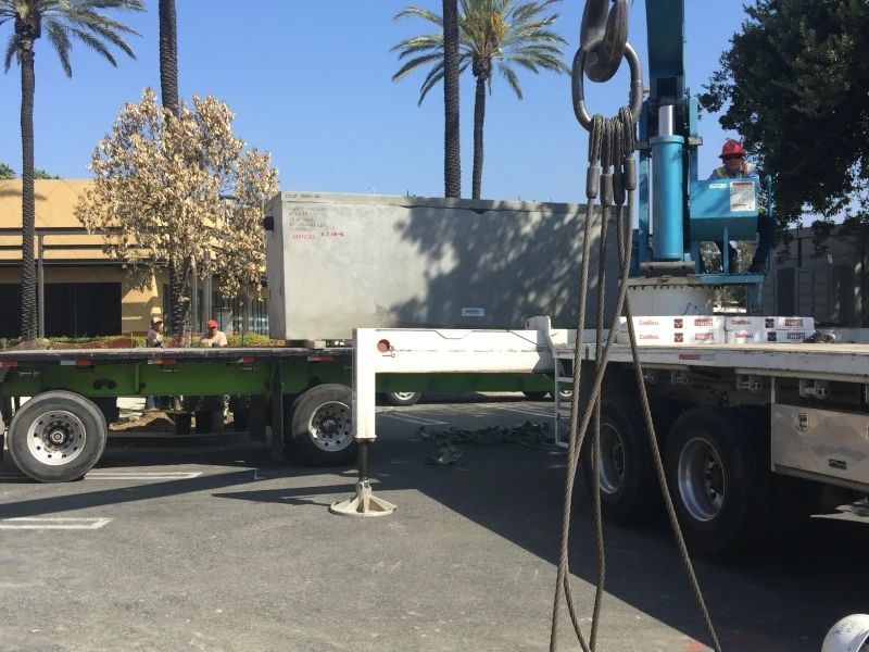 Crane lifting a concrete structure from a flatbed truck in a parking lot. Palms and buildings are in the background.