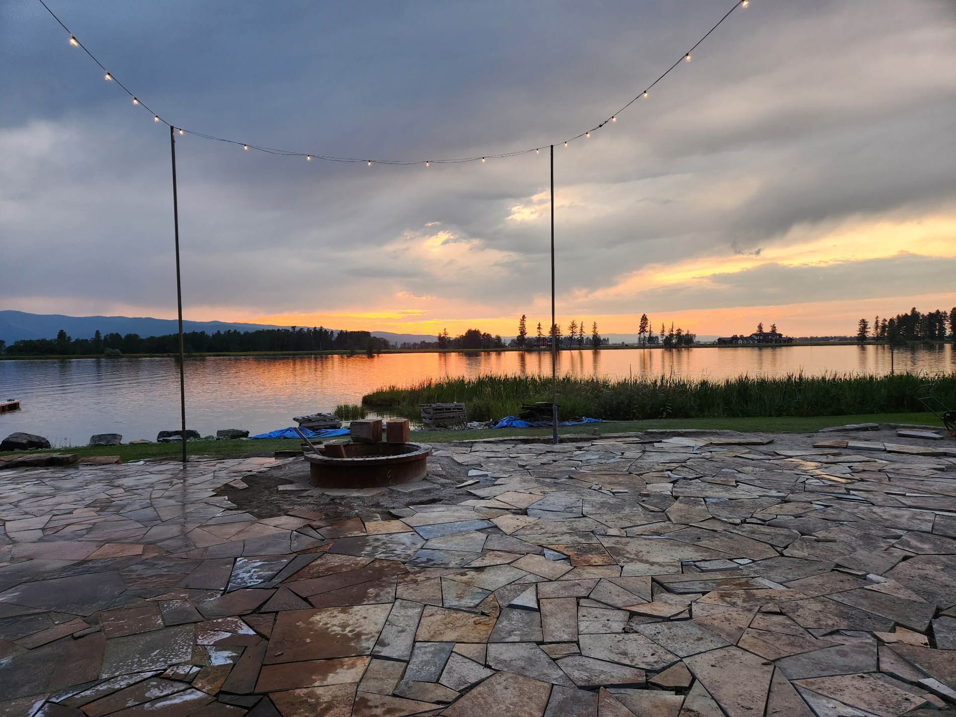 A patio with a fire pit and a view of a lake at sunset.