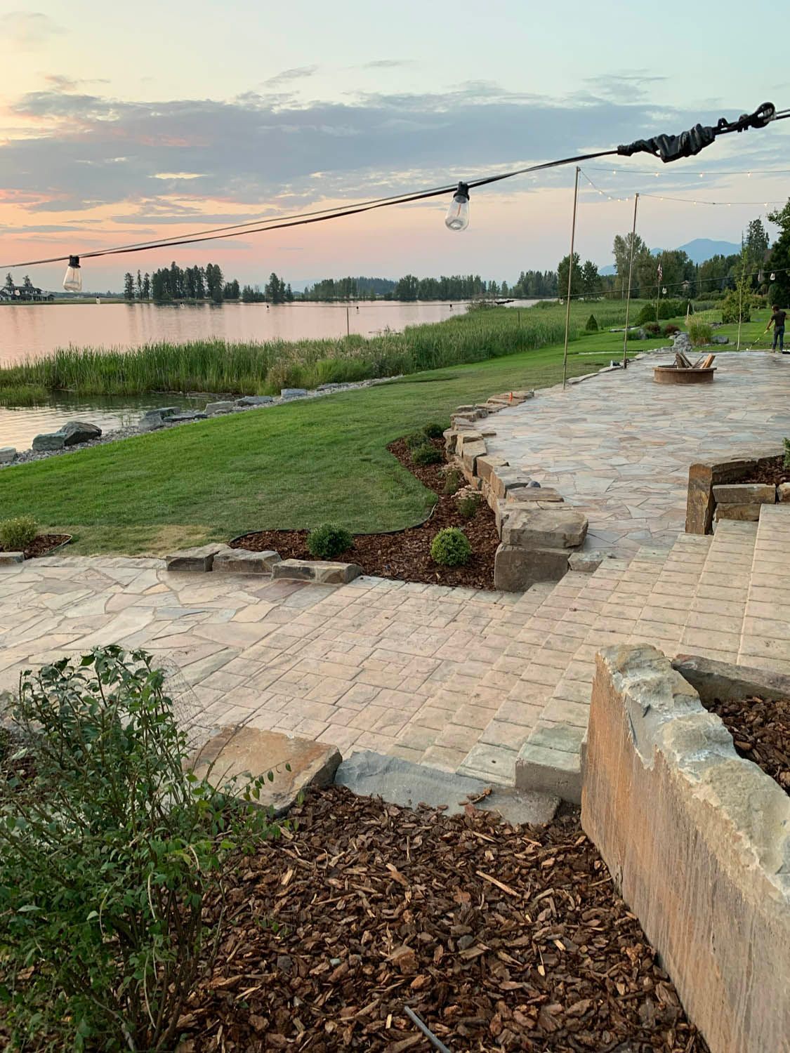 A patio with a fire pit and a view of a lake at sunset.