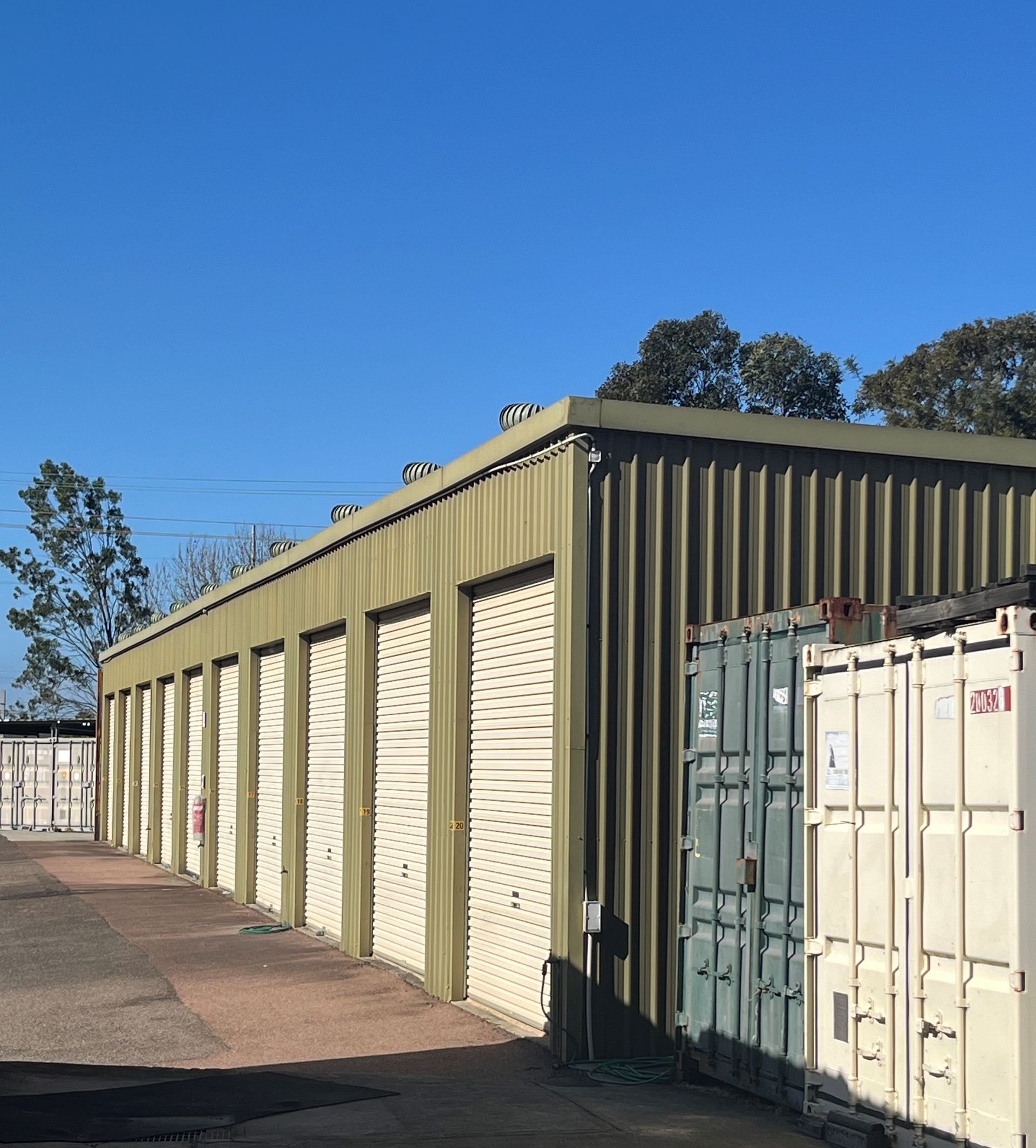 Side angle view of a row of yellow roll-up door storage units, with metal shipping containers in front and clear blue sky above. — Teralba Business & Storage Park in Teralba, NSW