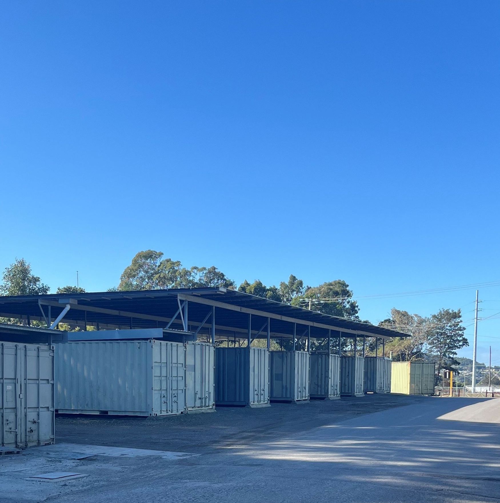 Shipping containers lined up beneath a long metal canopy beside a paved road, with tall trees and power lines in the background. — Teralba Business & Storage Park in Teralba, NSW