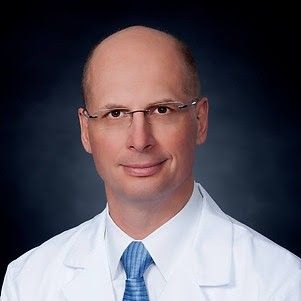 A man in a white lab coat and blue tie is smiling for the camera.