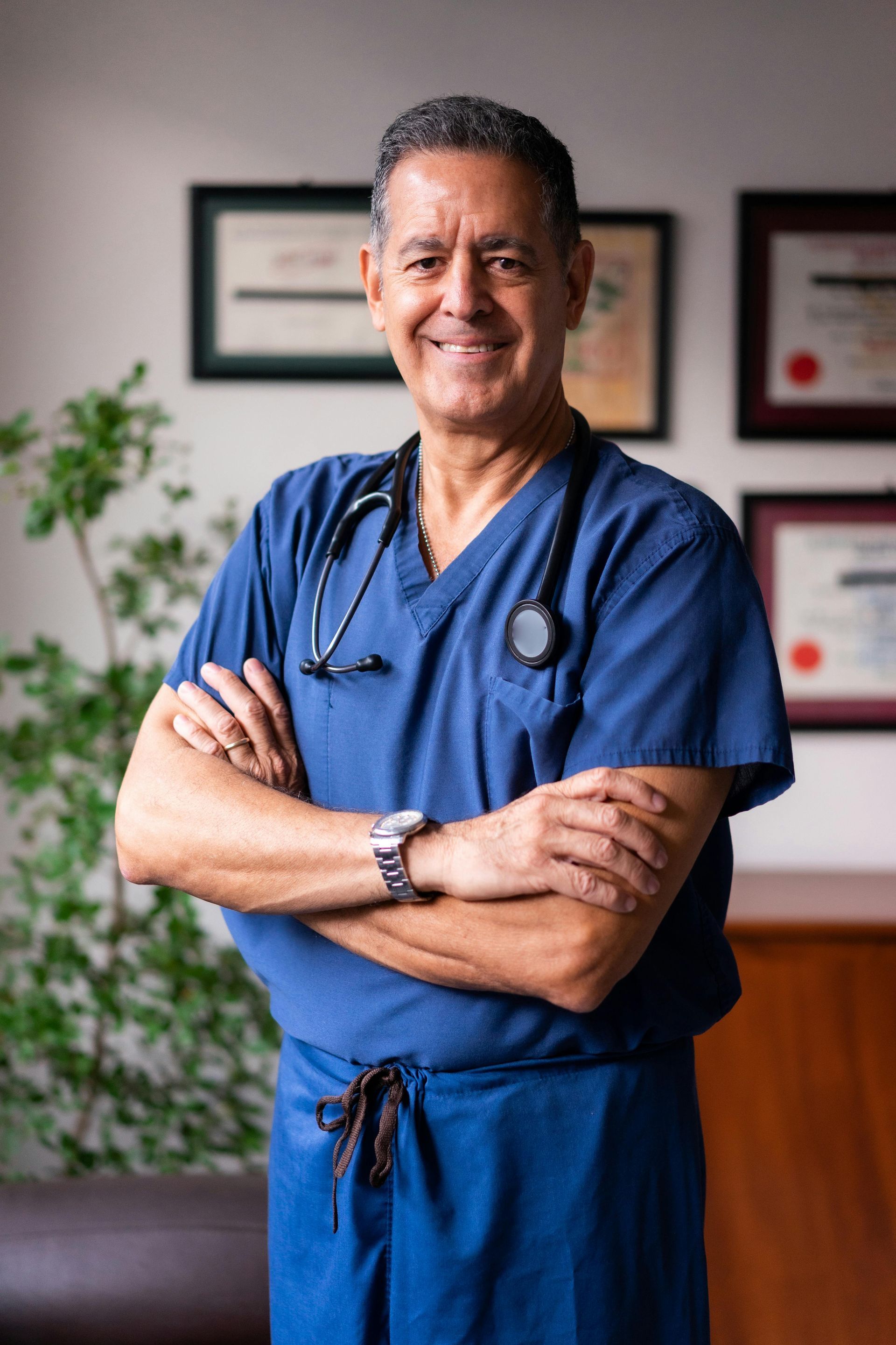 Doctor in blue scrubs standing with arms crossed in a clinic office, smiling at the camera.