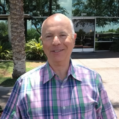 Bald man in pink, green, and blue plaid shirt smiles outdoors in front of a glass building.