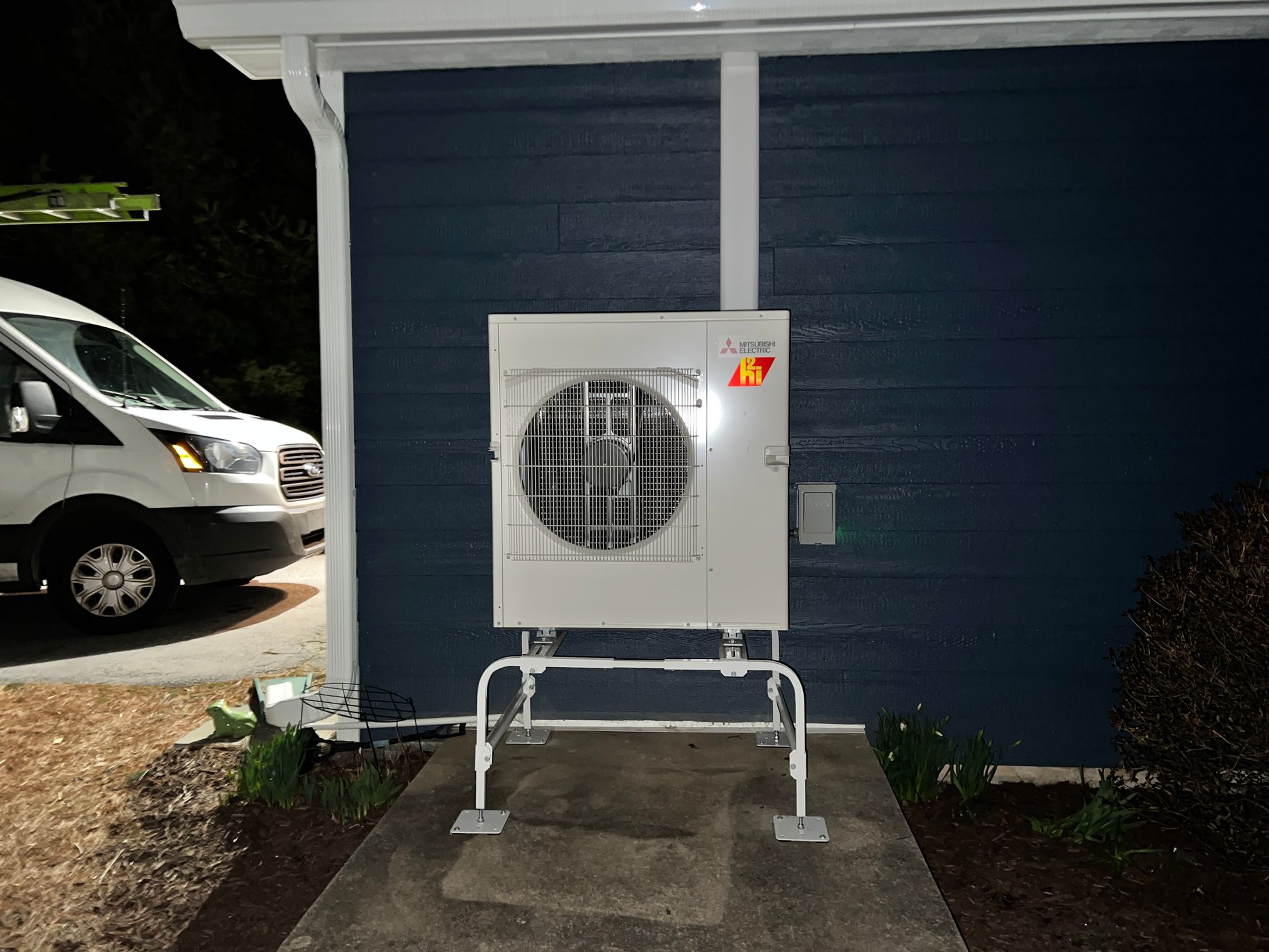 White heat pump unit on a metal stand mounted on blue building exterior with a white van in the background at night.