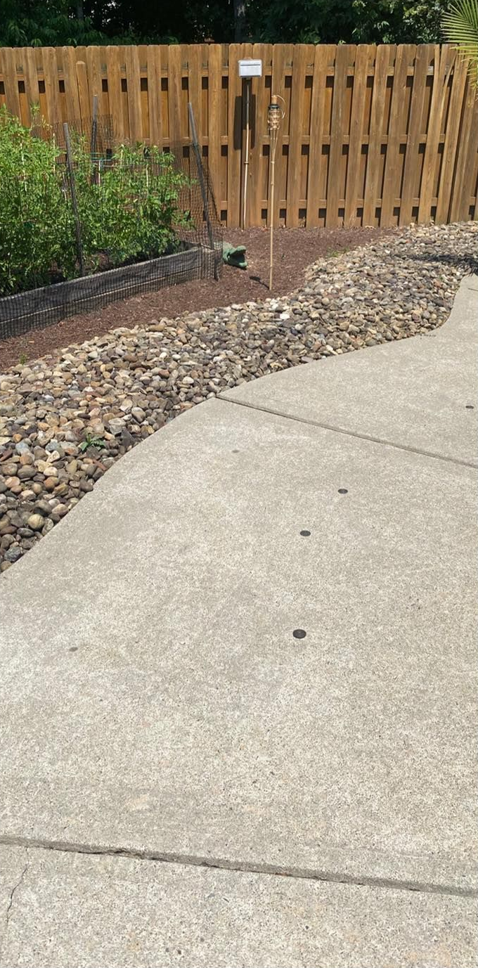 Concrete walkway borders a rock and mulch bed near a wooden fence.