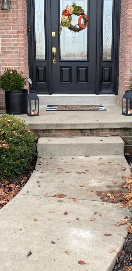 Front door with wreath and lanterns on the porch. Concrete walkway with leaves.