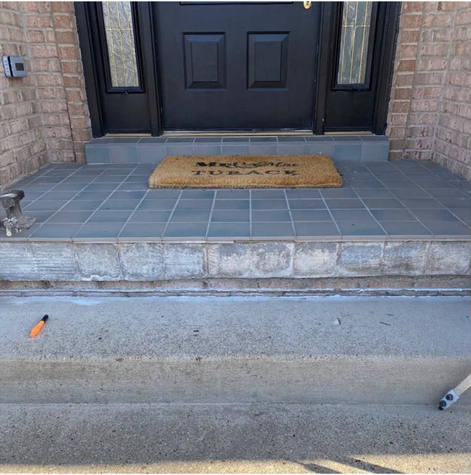 Front entrance with gray tiled porch and steps leading to a black door, doormat, brick wall.
