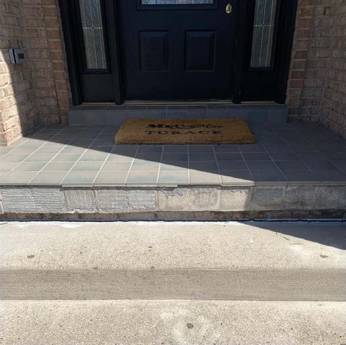 Exterior view of a home's entrance, with a concrete step, tiled porch, dark door, and welcome mat.