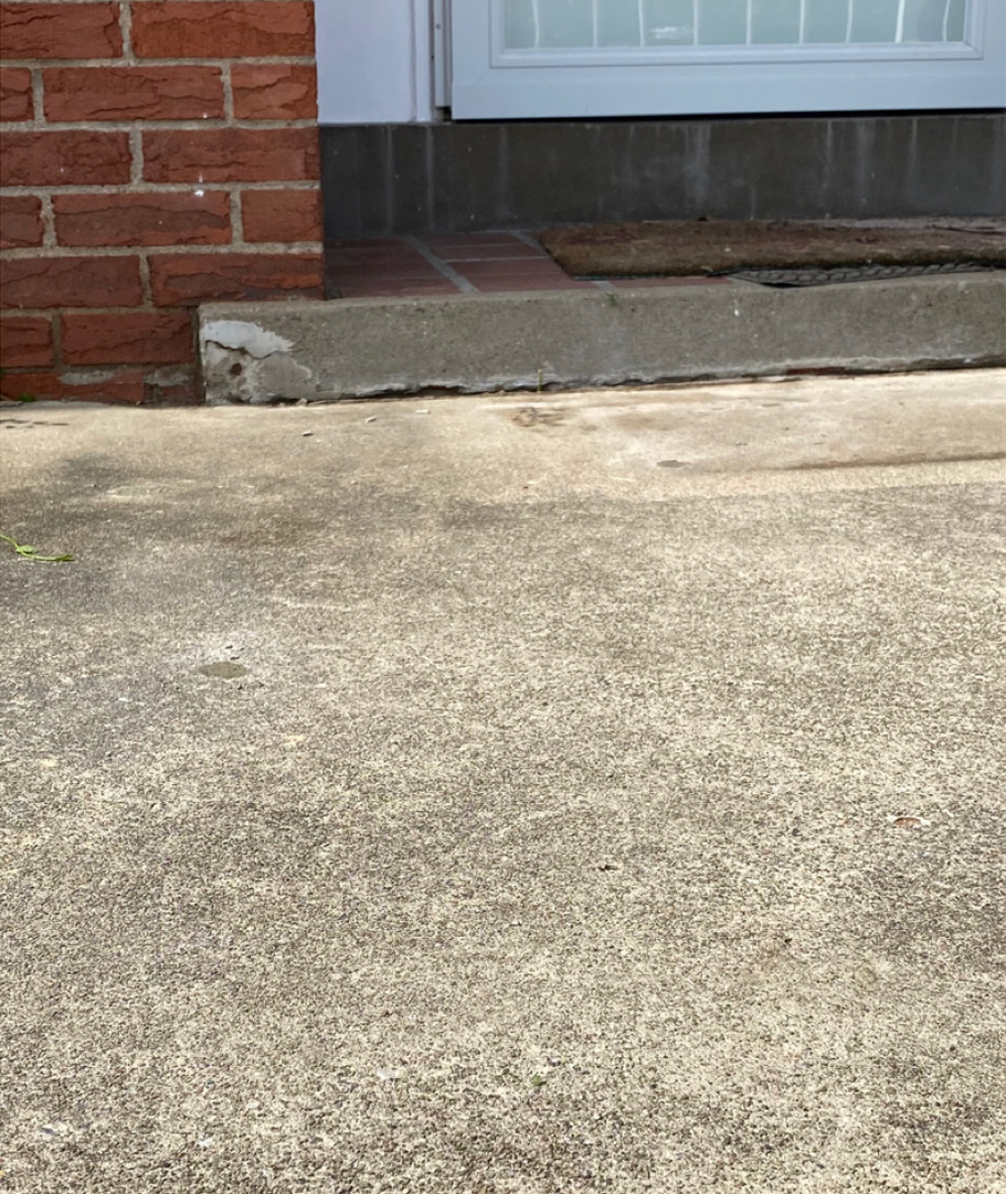 Brick building entrance with concrete step and a worn doormat on top of red brick tiles.