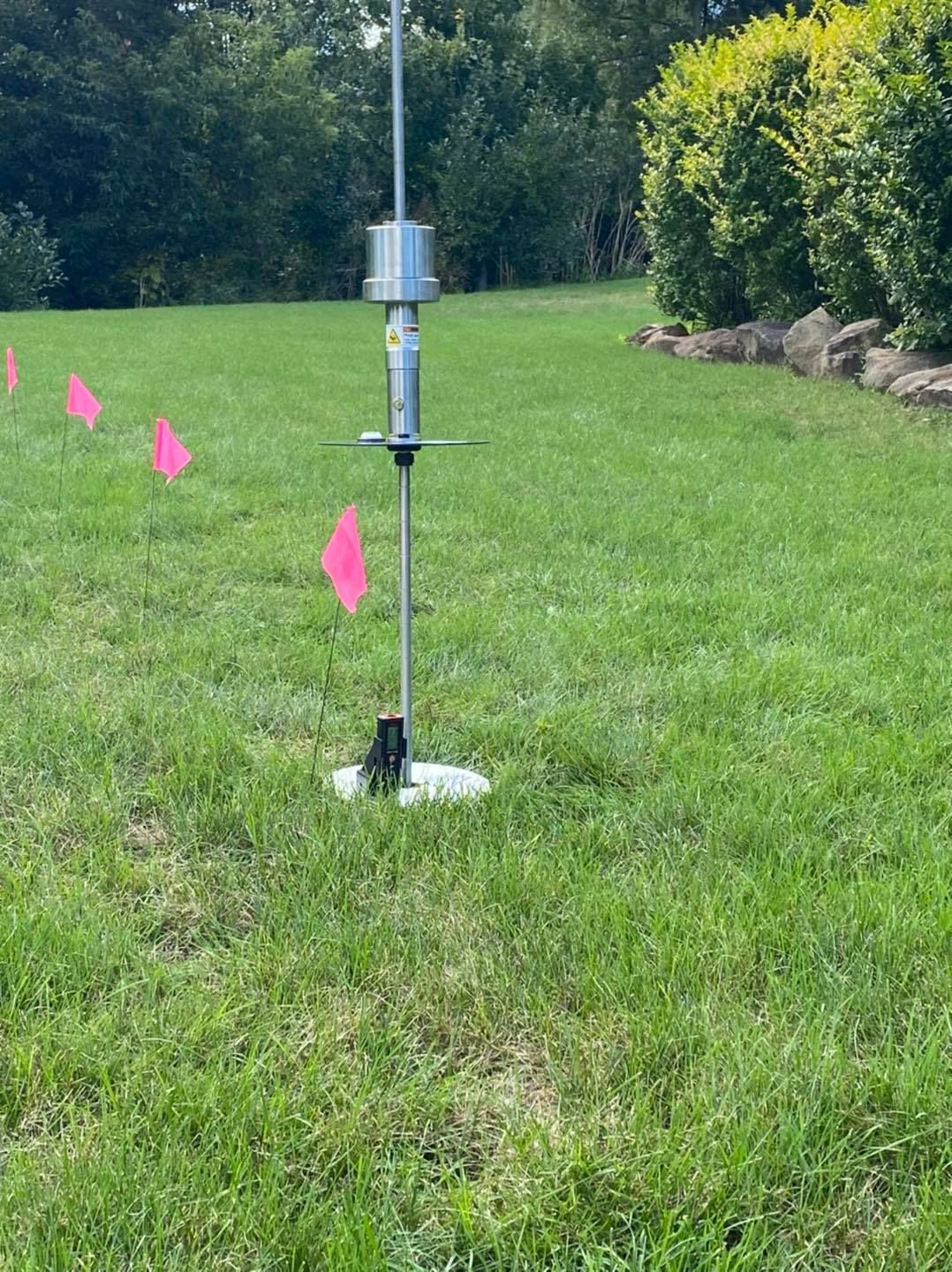 Tall metal device in grass, with pink flags. Green lawn, trees in background.