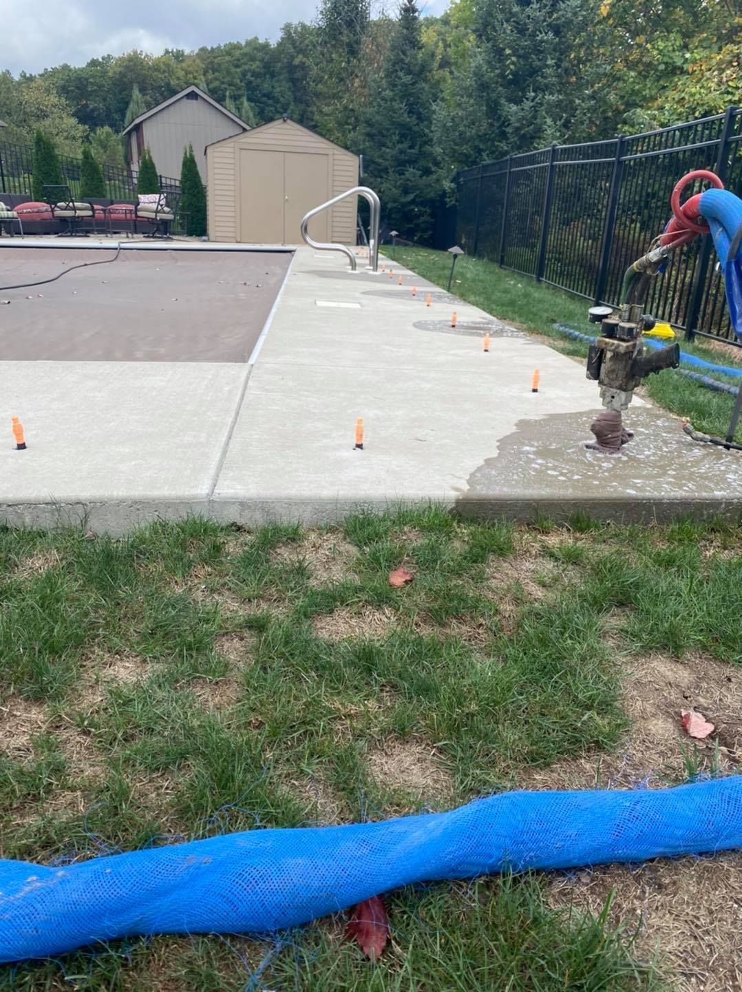 Concrete pool deck with orange markers; blue hose in foreground; a person by a water spigot.