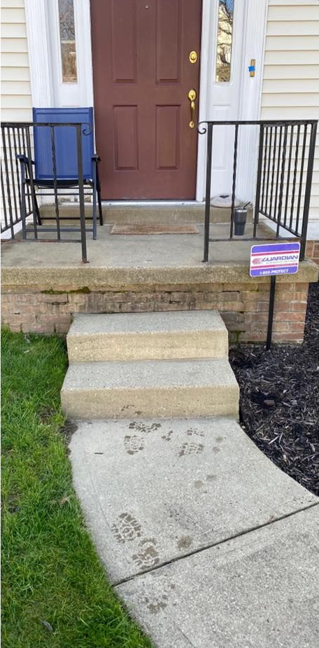Front entrance with concrete steps and walkway. Brown door, blue chair, black railing.