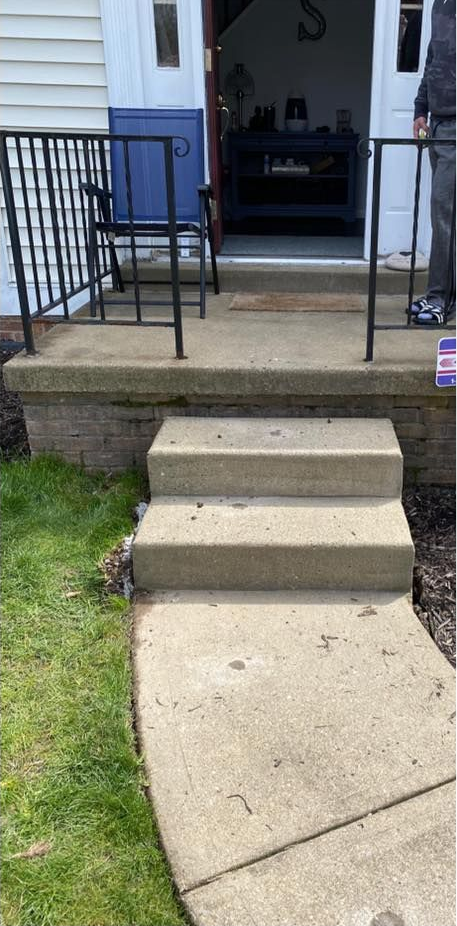 Concrete steps and walkway leading to a porch with black railing and blue chair.