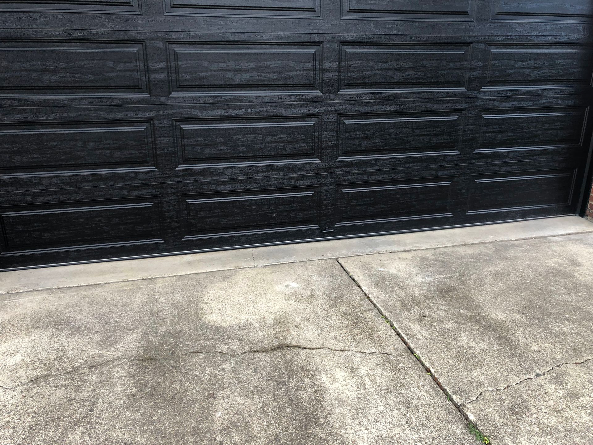Black garage door slightly ajar above cracked gray concrete driveway.