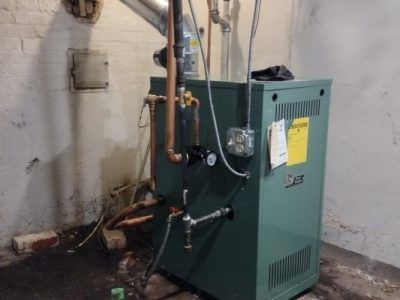 A green boiler is sitting in a basement next to a brick wall.