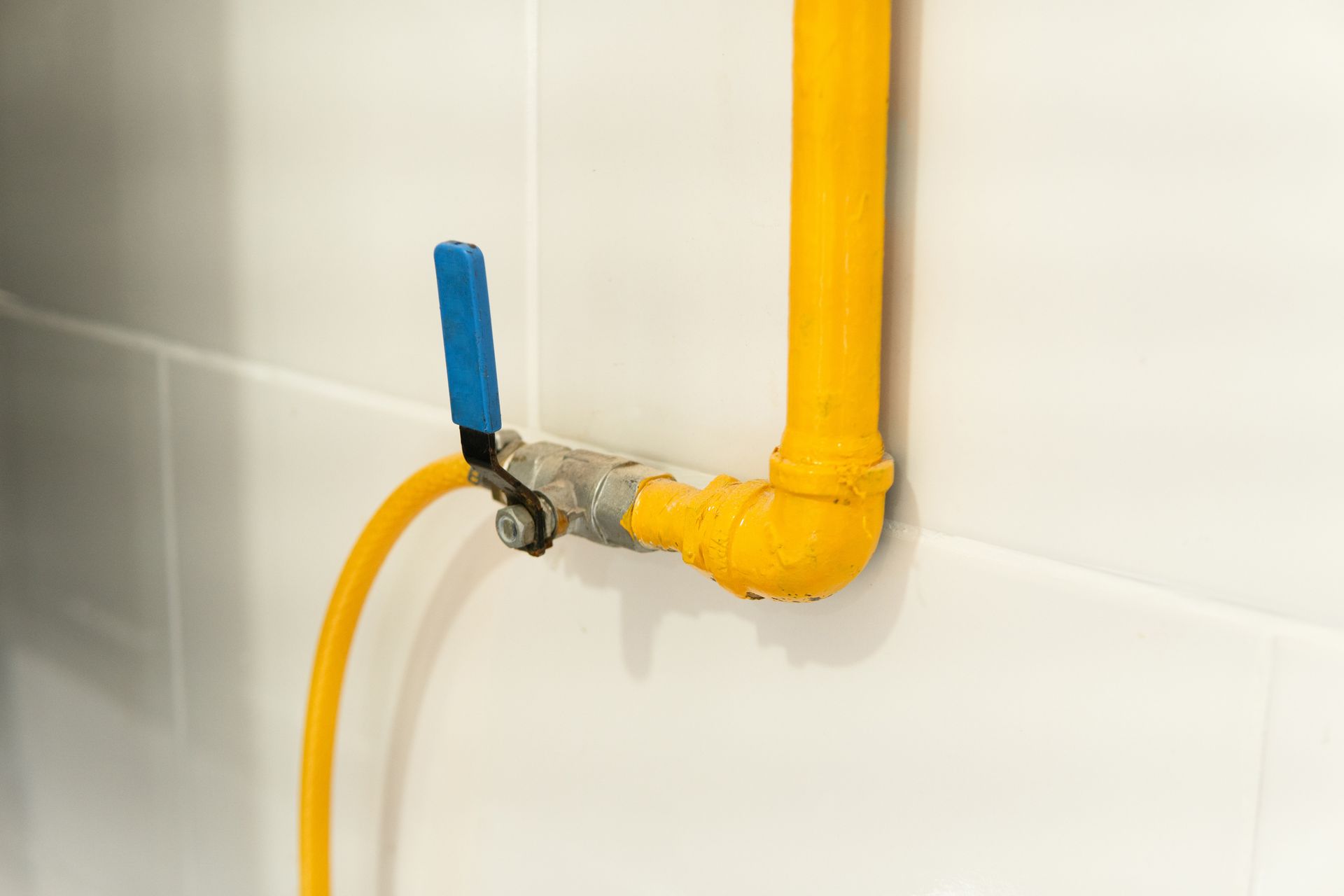 Yellow gas pipe with blue valve handle on white tiled wall.