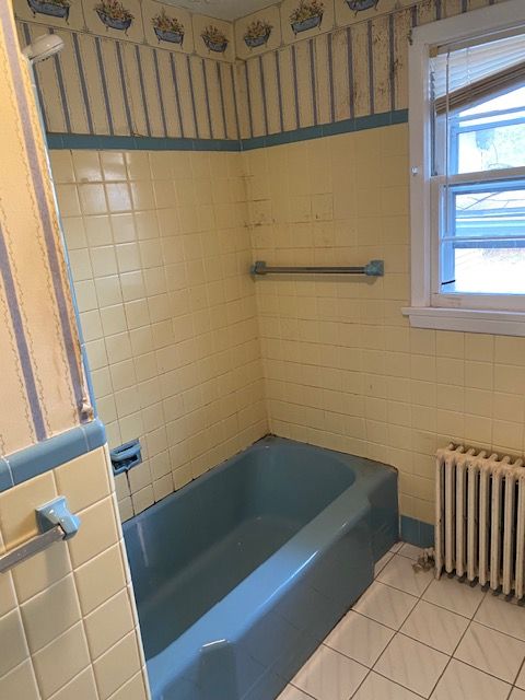 Blue and yellow tiled bathroom with a bathtub, window, and radiator.