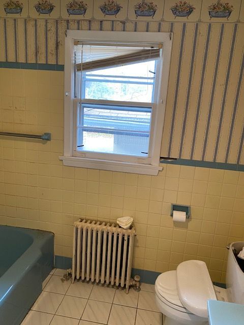 Bathroom with yellow tile, blue tub, white toilet, window, and radiator.