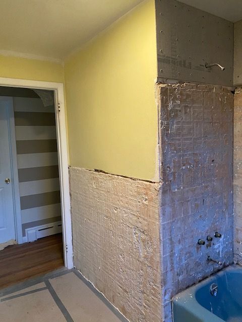 Bathroom renovation in progress; walls are partially stripped of tile and drywall. Yellow and gray surfaces. Blue bathtub.