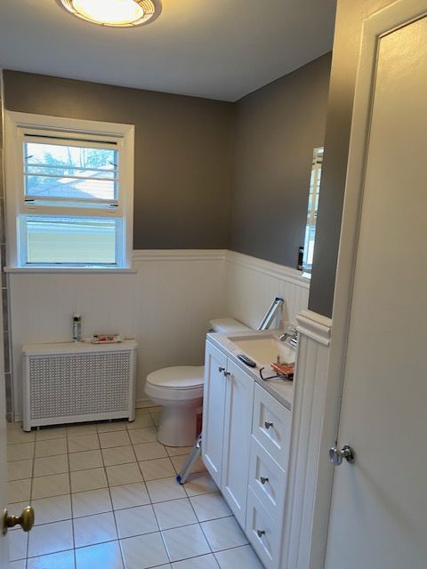 Bathroom with white vanity, toilet, and tile floor, gray walls above white wainscoting, and a window.