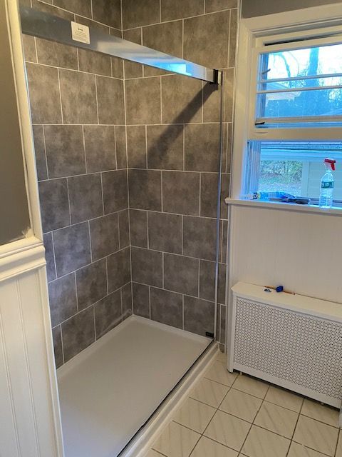 Gray tiled shower with glass door in bathroom, next to a window and radiator.