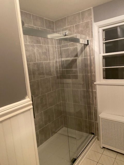 Gray tiled shower with glass door and white trim, next to a window and radiator.