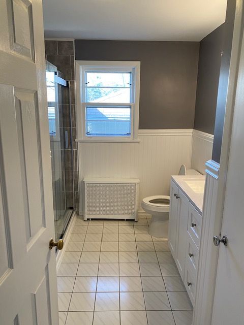 Bathroom with white wainscoting, gray wall, white vanity, shower, toilet, window, and a white radiator cover.