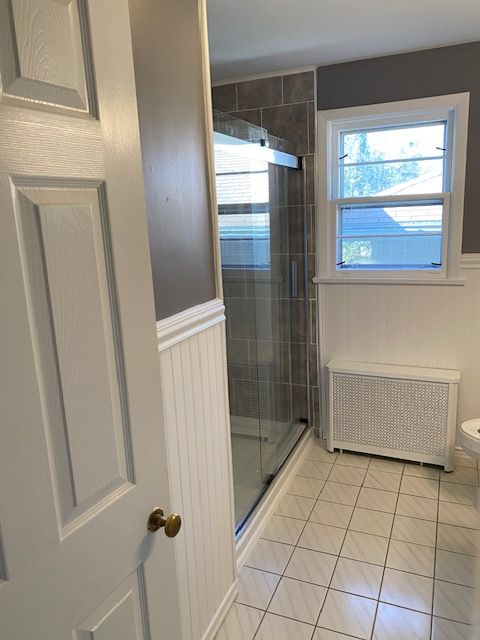 Bathroom with shower, window, and radiator. White door, tile floor, and gray walls.