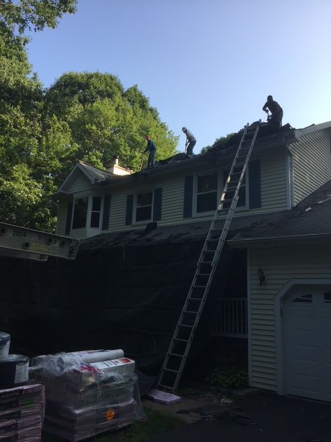 Roofers working on a house roof, using a tall ladder, with materials on the ground.