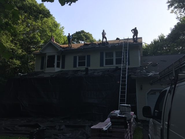 Roofers working on a house roof. A ladder is propped against the roof. Dark tarp covers the ground.