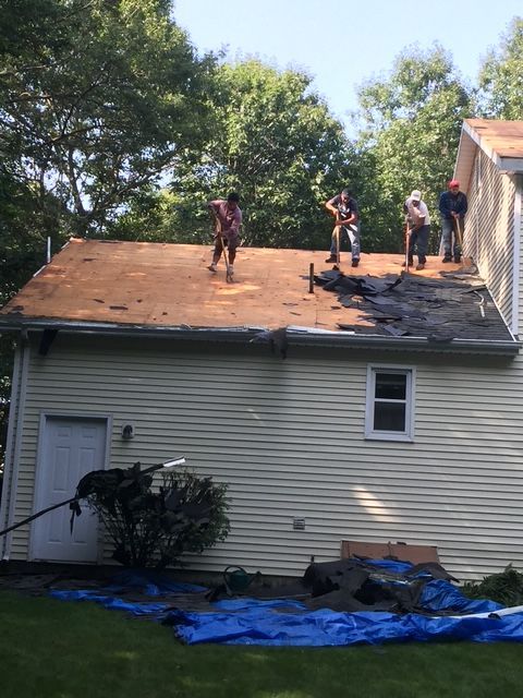 Roofers removing old shingles from a house with a green lawn and trees in the background.
